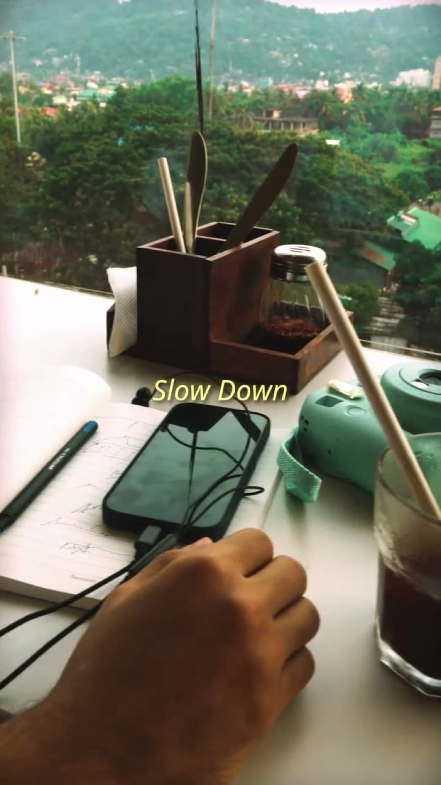 A notebook, phone and iced coffee on a window desk overlooking Guwahati's green hills