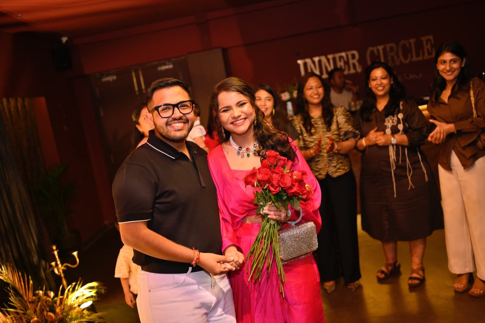 A couple with a bouquet of red roses, surrounded by friends