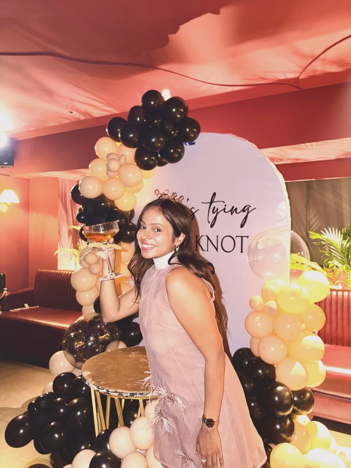 A bride-to-be beside a black and gold balloon arch that reads tying the knot